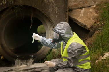 Bilim adamı koruyucu bir giysi, maske ve eldiven giymiş olarak nehirden bir örnek için sıvıyı aldı ve kanalizasyon borusuyla onun rengini karşılaştırdı.