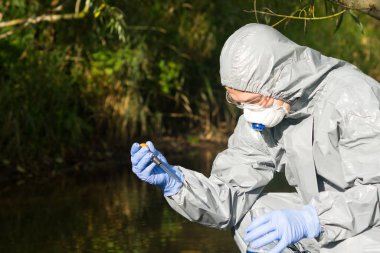 Koruyucu giysili ve maskeli bir adam elinde nehirden bir pipet su tutuyor ve suyun kalitesini incelemek için test tüpüne döküyor.