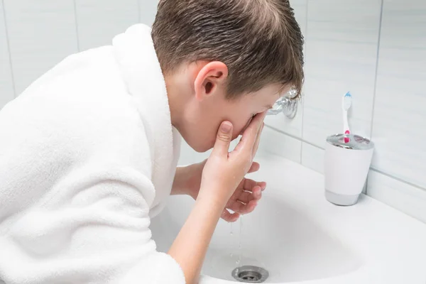 a child in a white bathrobe washes his eyes with water in the bathroom