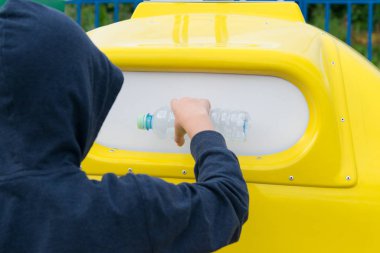 Mavi kapüşonlu bir çocuk çöp kutusuna boş bir şişe atar.