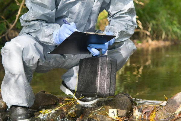 the specialist, in a protective suit and with a suitcase in his hand, is recording the findings at the crime scene on the pond