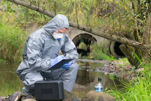 a specialist, in a protective suit and gloves, working with toxic substances, records the results of a sample of liquid from the river, on a sheet on a tablet, against the background of cleaning pipes