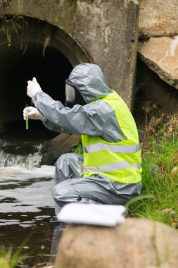 Açık hava kazasından sonra elinde test tüpü ve üzerinde test suyu olan bir pipet tutan koruyucu kıyafetli bir adamın yan görüntüsü.
