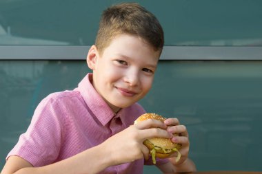 Çocuk elinde bir hamburger tutuyor ve yemeden önce gülümsüyor.