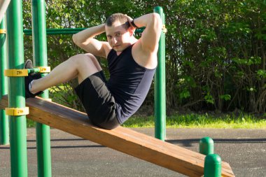 Spor sahasında bir adam karın kasları üzerinde egzersiz yapar ve kameraya bakar.