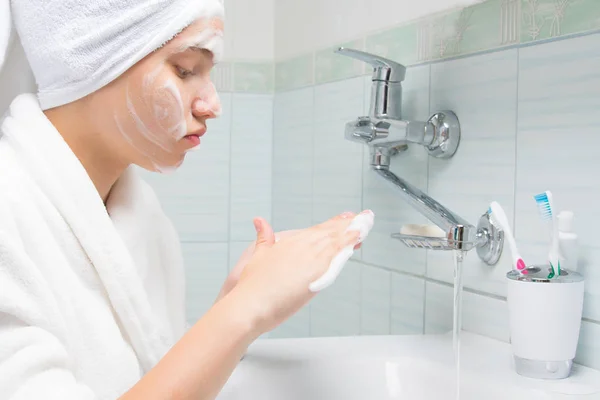 a girl with a white towel on her head and in a dressing gown, in the bathroom, washes foam, for cleansing, from her face, near a tap with water