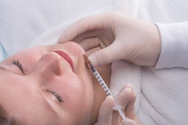Steril eldivenli doktor, çenesinin bir kısmında bir kadına güzellik iğnesi yapıyor.
