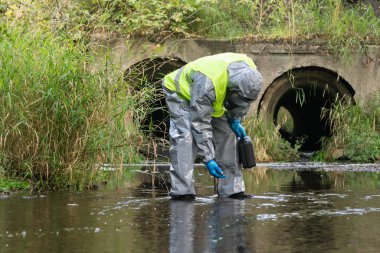 Koruyucu giysi giyen bir uzman elinde ekipmanla bir bavul tutuyor ve nehirdeki su kirliliğini özel bir aletle ölçüyor.