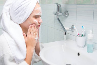 Beyaz önlüklü kız, yüzünü köpükle temizliyor, banyoda, musluğun yanında ve batıyor.