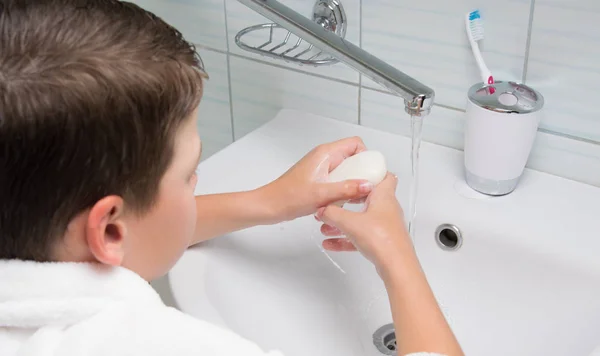 boy, in a white bathrobe, in the bathroom, washes his hands with soap, over the sink and faucet