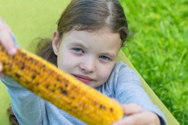 Kız yeşil çimlere oturur ve güzel sarı bir mısır koçanı gösterir.