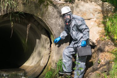 Koruyucu giysi ve maske giyen bir çevresel laboratuvar uzmanı elinde gezici bir laboratuvar olan bir çantayı tutuyor ve kimyasal akışının merkezine gidiyor.