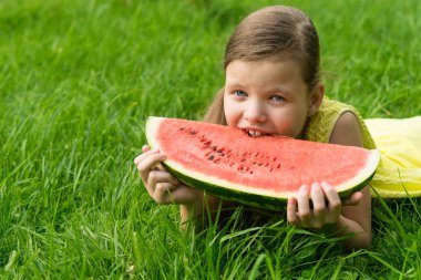 Kız büyük lezzetli bir karpuz dilimini ısırıyor, yeşil çimlerin üzerinde yatıyor.