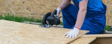 Mavi üniformalı ve koruyucu eldivenli bir işçi elektrikli testereyle ahşap bir kalkanı kesiyor.