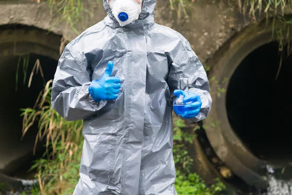 a specialist in protective clothing and a mask holds in one hand a flask with a reagent and shows the thumb up sign with the other hand
