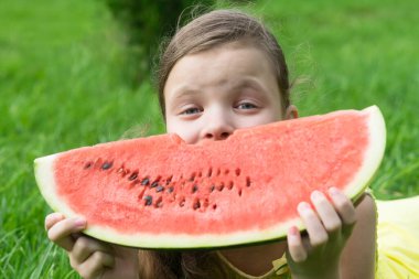 Kız büyük kırmızı bir karpuz diliminin arkasına saklanmış çimlerin üzerinde oturuyor.
