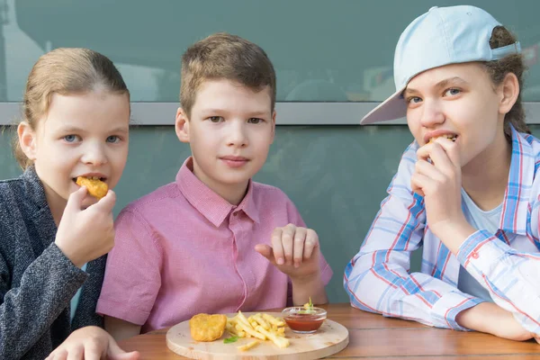 小学生の男の子と2人の女の子がテーブルに座ってファーストフードを食べます — ストック写真