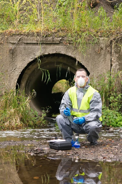 a man in a protective suit and a respirator against bacteria and viruses sits by the river next to a mobile laboratory for research