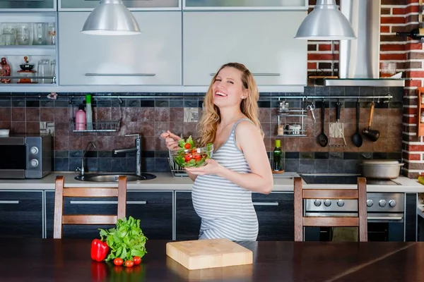 Gülümseyen hamile kadın mutfakta sebze salatası yiyor.