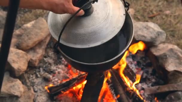 Turista levanta a tampa para olhar ou água fervente na panela. Feche a mão humana. Cozinhar em uma panela na fogueira . — Vídeo de Stock