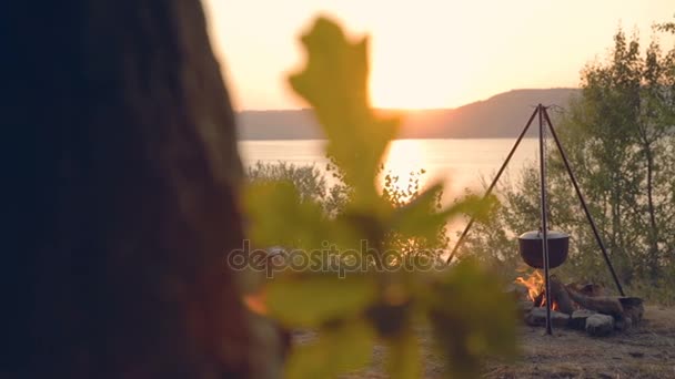 DOLLY MOTION: Cocinar en llamas para la compañía al atardecer. En una olla de hierro fundido sobre un fuego en el lago. Preparación para cocinar al aire libre . — Vídeo de stock