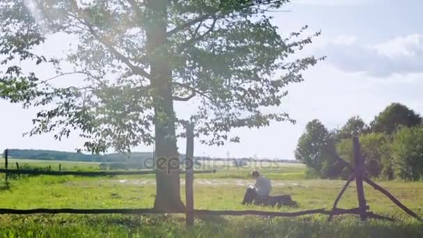 Een kleine getalenteerde jongen zitten onder een boom achter een houten hek en een landschap schilderen. Het landschap is bij de zonsondergang. Slow motion. — Stockvideo