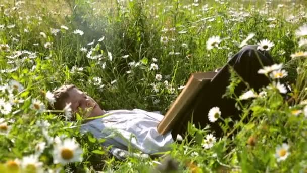 Маленький деревенский парень лежит в высокой траве и читает старую книгу — стоковое видео