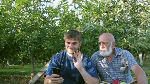 Los agricultores en el jardín con un teléfono inteligente hablan a través de la comunicación por vídeo. Brazo oscilante y sonrisa — Vídeos de Stock