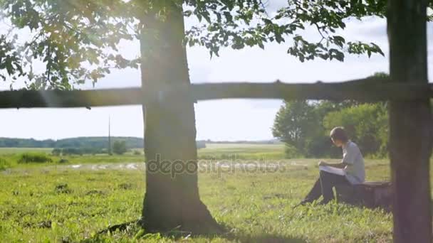 Хлопчик сидить біля дерева і малює олівець на папері. Промені сонця через листя дерев. Повільний рух — стокове відео