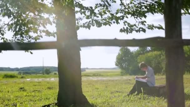 Молодий художник, сидячи під деревом, малює літній пейзаж — стокове відео