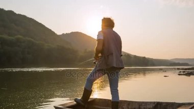 Genç adam gün batımında tekneyle gölde balık tutuyor.