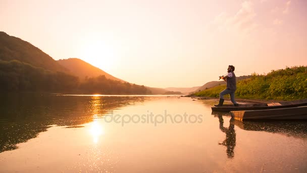 A cute teen catches a fish from a wooden boat on the river. The sun is hiding behind the mountain — Stock Video