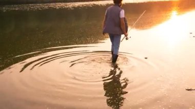 Genç balıkçı günbatımı için gider. Lastik çizmeler giyen su geçiyor. Balıkçı siluet su görünür