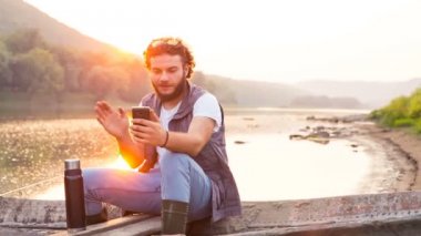 Şık güzel balık adam ahşap bir tekne bir smartphone cep telefonu ile konuşmak. Gün batımında Skype konuşuyor.