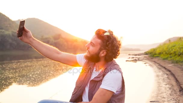 Un joven pescador elegante se sienta en un barco de madera y hace un selfie divertido contra el telón de fondo de la puesta del sol y el río — Vídeos de Stock