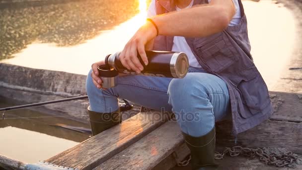 Un joven pescador vierte un café sentado en un bote de madera. El pescador hipster está tomando café al atardecer. — Vídeos de Stock