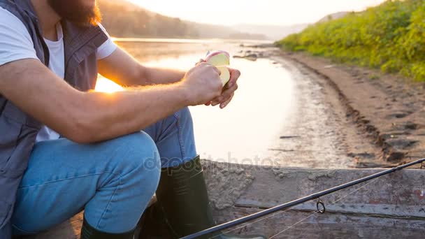 Un joven pescador come una manzana sentada en un barco pesquero en el río. Noche cálida sol-verano — Vídeos de Stock