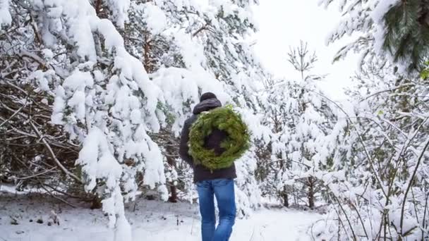 Um homem usa uma coroa de Natal numa floresta coberta de neve, atirando-a para o ombro — Vídeo de Stock
