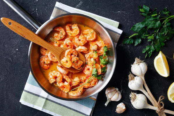 Shrimp scampi with garlic butter sauce served with lemon and sprinkled by parsley, on a frying pan with ingredients on a concrete background, view from above