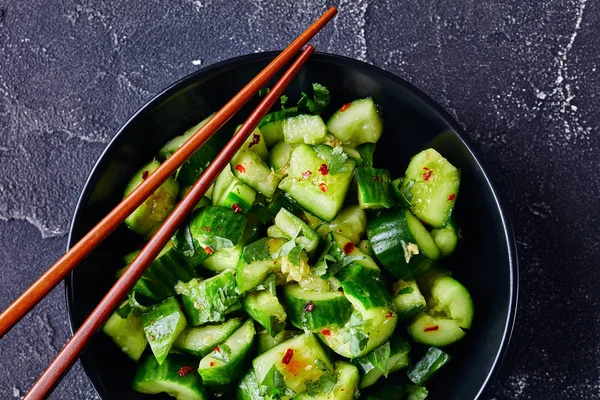 pai huang gua, Chinese Smashed Cucumber salad sprinkled with fresh coriander leaves in a black bowl with chopsticks on a concrete table, flat lay, close-up, macro