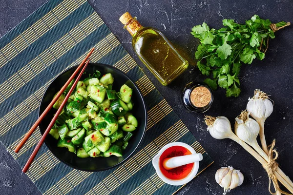 Chinese Smashed Cucumber salad, pai huang gua, with chili sesame oil garlic soy sauce dressing sprinkled with fresh coriander leaves in a black bowl on a concrete table with ingredients, flat lay