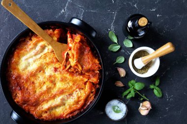 Traditional italian cannelloni pasta stuffed with bolognese sauce and mozzarella cheese on top on a black baking dish on dark concrete background with fresh basil, sea salt, and garlic, top view clipart