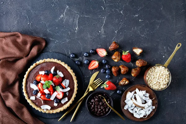 Sugar-free healthy dessert: chocolate tart with fresh strawberries, blueberries, coconut chips, served on a dark concrete background with ingredients: dates, rolled oats, top view, copy space