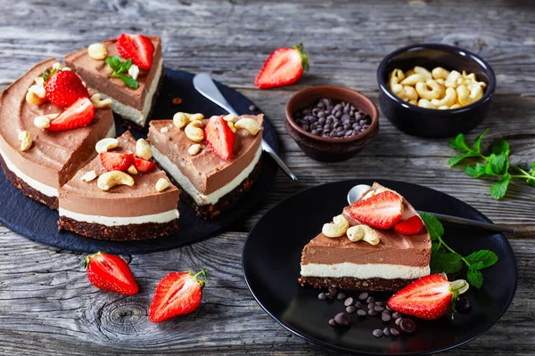 Raw triple chocolate cashew tart made of coconut cream and soaked cashews with the crust of raw dates, cocoa, and nuts on a black plate with strawberries and mint on a wooden background, close-up