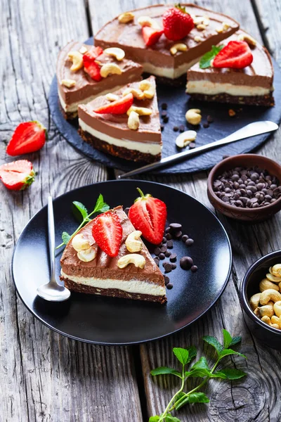 Triple chocolate cashew tart made of coconut cream and soaked cashews with the crust of raw dates, cocoa, and nuts on a black plate with strawberries and mint on a wooden background, close-up