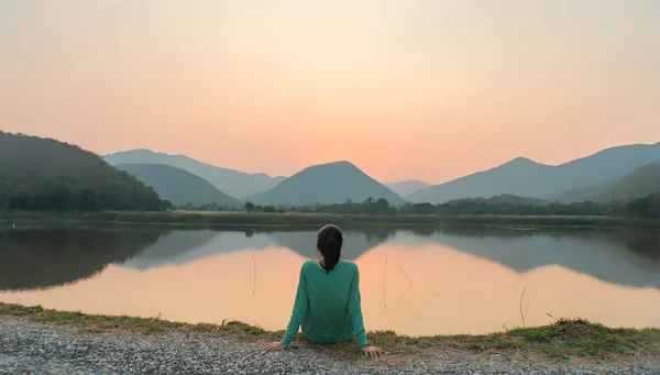 A woman is sitting by water and staring at sunset silently.