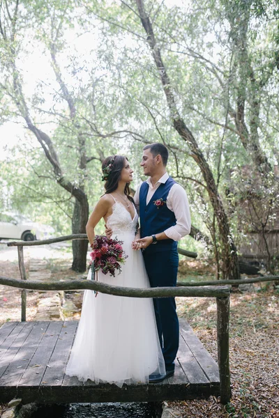 Noivo feliz em um terno azul escuro abraça uma noiva bonita em um vestido de noiva branco ao ar livre em uma ponte de madeira — Fotografia de Stock