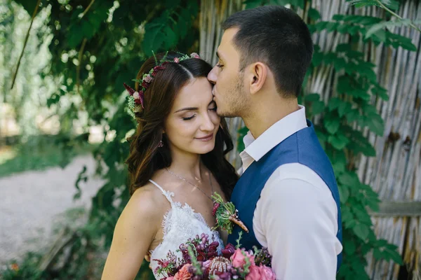 Happy groom in a dark blue suit kisses a beautiful bride in a white wedding dress with pink and purple bouquet of flowers outdoors — Stock Photo, Image