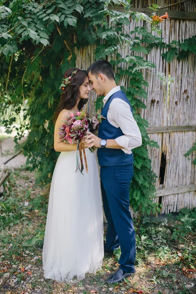 Felice sposo in un abito blu scuro abbraccia e tiene la mano della bella sposa in un abito da sposa bianco con mazzo di fiori rosa e viola all'aperto — Foto Stock