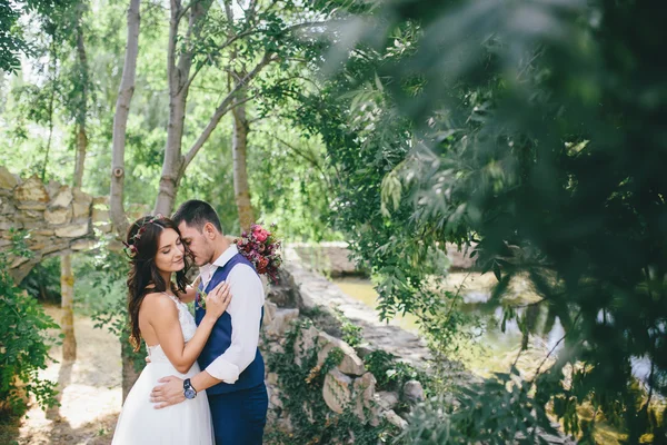 Happy groom in a dark blue suit hugs a beautiful bride in a white wedding dress with pink and purple bouquet of flowers outdoors on the background of a stone arch — Stock Photo, Image